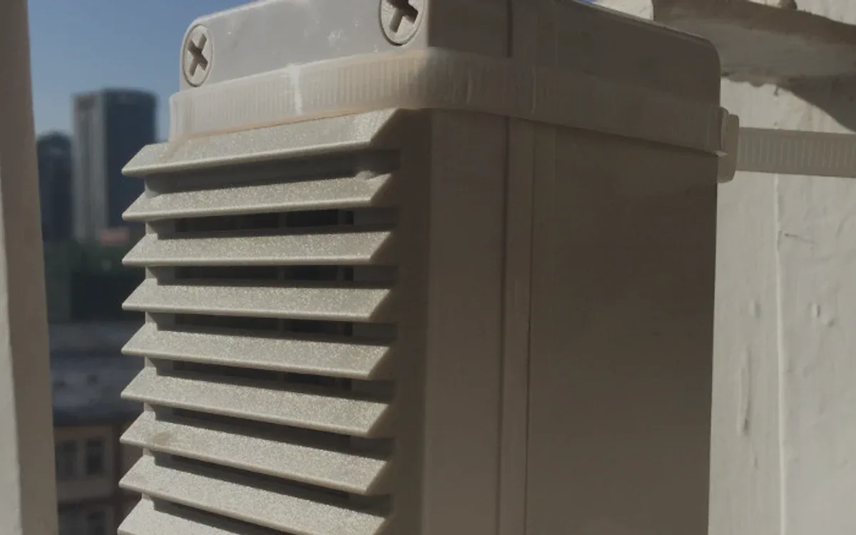 Nodo de sensores instalado a la intemperie: la caja con su abrigo de lamas, sujeta con bridas a un soporte de pared, con la ciudad al fondo